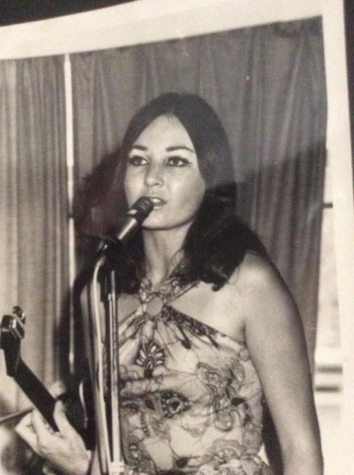A black and white print of a young Ann with long dark hair, singing into a microphone on a stand in a patterned halter-neck dress, with a curtain behind her.
